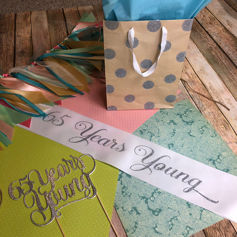 65 years young silver glitter cake topper shown with birthday sash.