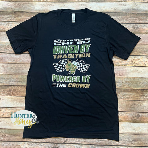A black t-shirt on a wood-planked background. The t-shirt has a 3-color glitter design on the front (in old gold, white, and dark green). The design reads: "Damascus Cheer: Driven by Tradition, Powered by the Crown." The design features a hornet wearing a crown flanked by two racing checkered flags on either side. The tee is a crewneck Bella + Canvas unisex, tri-blend short-sleeve tshirt.