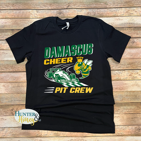 A black t-shirt with a 3-color graphic that reads Damascus Cheer Pit Crew and has a race car crossing the checkered finish line with a hornet wearing a crown. The design is in dark green, gold, and white smooth heat transfer vinyl. There is Hunter & Honey logo in the lower left corner.
