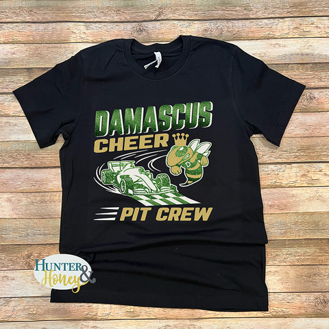 A black t-shirt with a 3-color graphic that reads Damascus Cheer Pit Crew and has a race car crossing the checkered finish line with a hornet wearing a crown. The design is in dark green, gold, and white glitter. There is Hunter & Honey logo in the lower left corner.