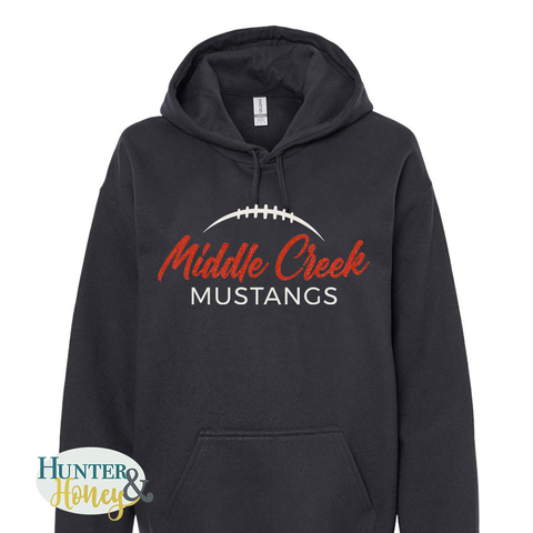 A black pullover hoodie with a front kangaroo pocket and a graphic print reading 'Middle Creek MUSTANGS' in red and white glitter, featuring a football design above the text. The hoodie is shown on a plain white background with a Hunter Honey & logo watermark in the bottom-left corner.