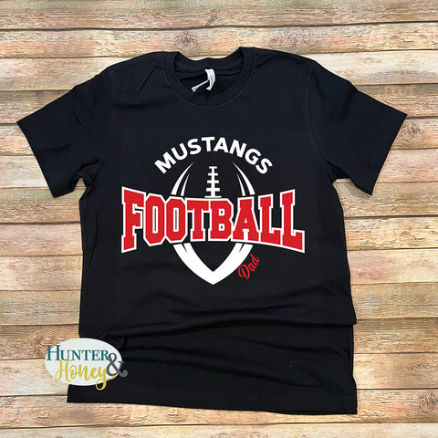 A black short-sleeve t-shirt with a 'Mustangs Football Dad' graphic, laid flat on a rustic wooden plank background. The graphic features a white football silhouette and the words 'MUSTANGS FOOTBALL' in white and red, with 'Dad' written in red script.