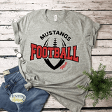 A heather gray graphic t-shirt featuring the words 'MUSTANGS FOOTBALL MOM' in black and red, with a football graphic in the background. The shirt is styled in a flat-lay arrangement with blue jeans, a watch, and a small green plant on a white wood plank background.