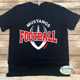 A black short-sleeve t-shirt with a 'Mustangs Football Mom' graphic, laid flat on a light brown wooden plank background. The graphic features a white football outline, red 'FOOTBALL' text, and white 'MUSTANGS' text.