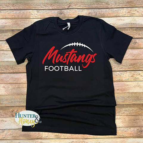 A black short-sleeve t-shirt with a 'Mustangs Football' graphic printed on the front, laid flat on a rustic wooden plank background. The graphic features 'Mustangs' in red script, 'FOOTBALL' in white block letters, and a white football graphic arching above the text.