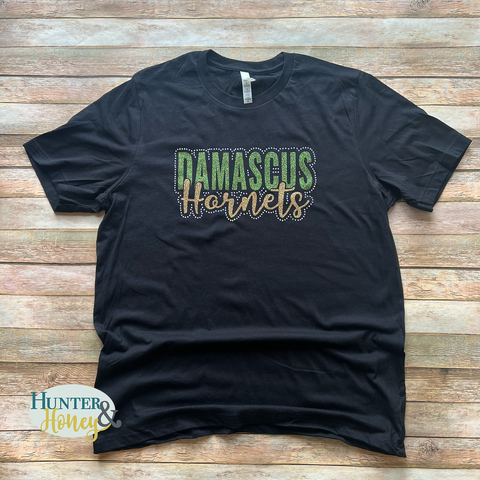 A photo of a black cotton crew neck short sleeve t-shirt with a Damascus Hornets logo in dark green and gold glitter surrounded by AB rhinestones. 